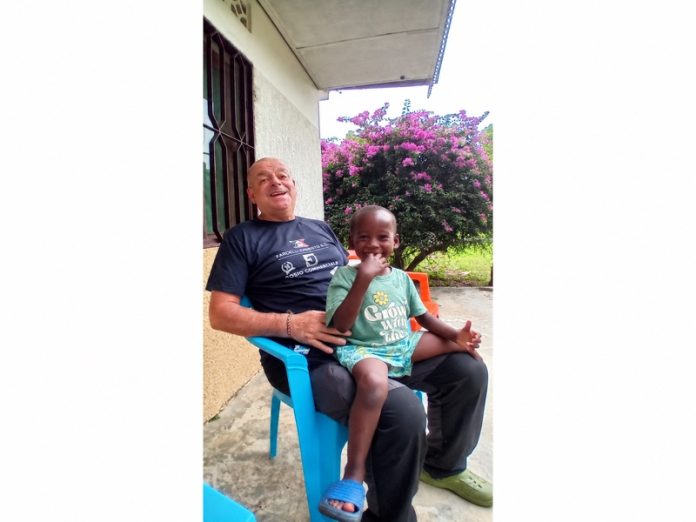 Un uomo e un bambino sorridenti, seduti all’aperto accanto a un arbusto fiorito a Lusanga.