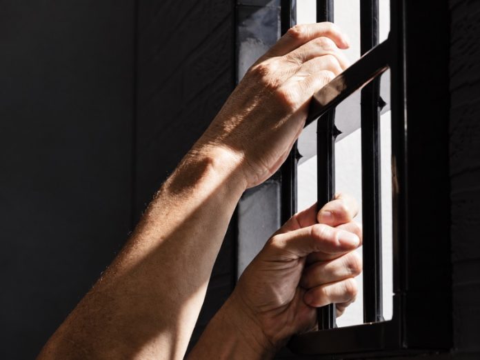 Due mani afferrano le sbarre di una finestra illuminata dalla luce esterna.